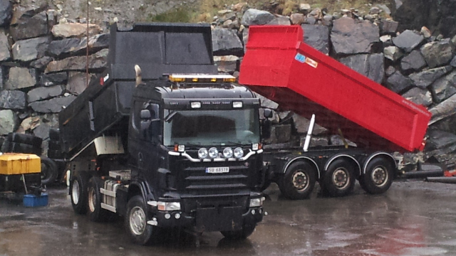 En svart lastebil med tilhenger som tømmer en rød container på en steinete byggeplass.