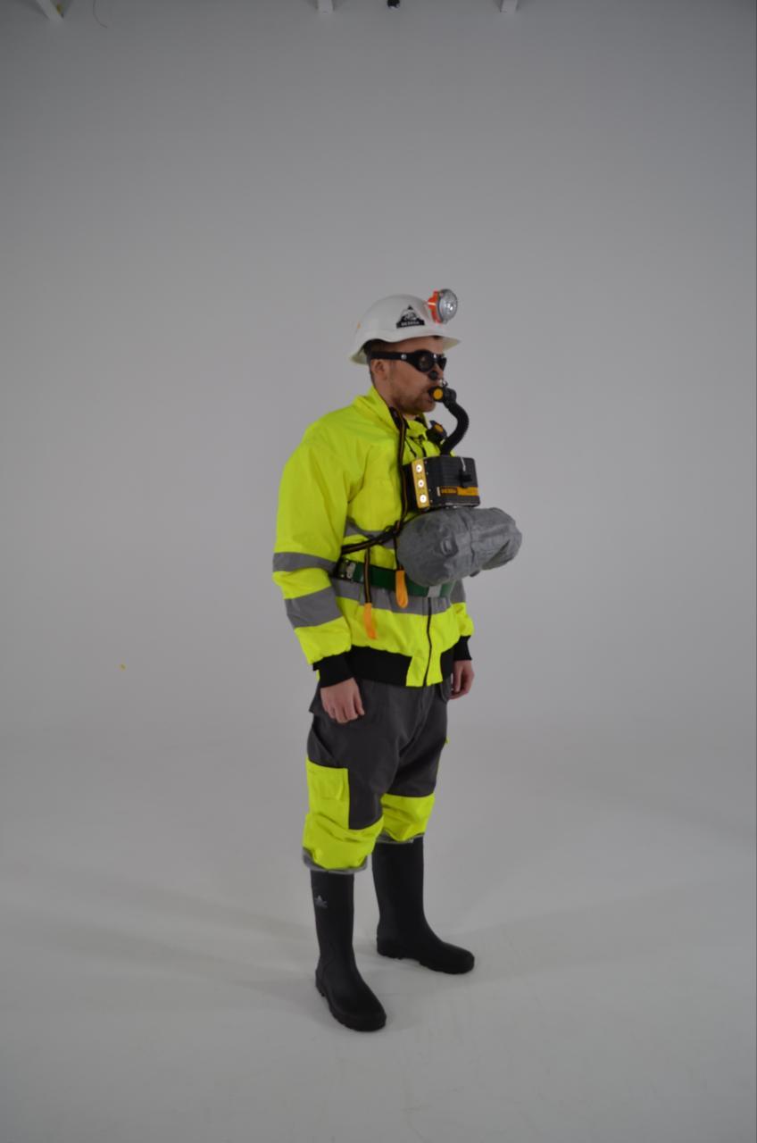 En mann i verneutstyr med hjelm og gassmaske, iført refleksjakke og støvler, står på en lys bakgrunn.
