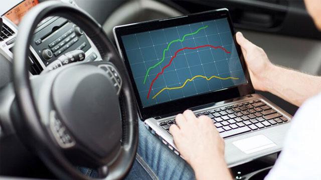 En person sitter i en bil og bruker en laptop som viser grafer med grønne og gule linjer.