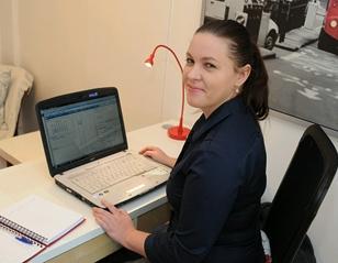 Kvinne som sitter ved et skrivebord med en bærbar datamaskin og notatblokk, smilende mot kameraet.