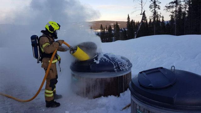 Brannmann som bruker en brannslange for å slukke en brann i en snødekt hage med søppeldunker i bakgrunnen.