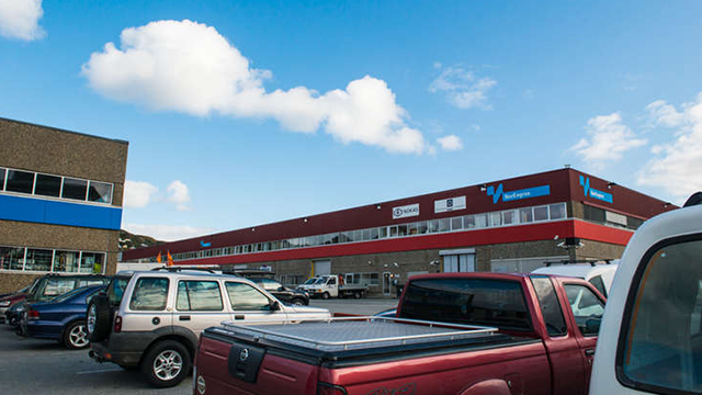 En parkeringsplass med flere biler foran en stor bygning med blå og rød fasade under en klar himmel.