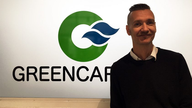 En mann med skjegg står foran en vegg med "GREENCAP" logoen og bølger i grønt og blått.