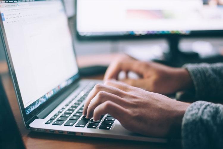 En person skriver på en laptop med to dataskjermer i bakgrunnen.