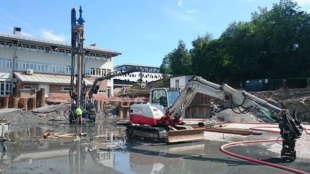 Byggeplass med gravemaskiner og boreutstyr som arbeider på en grunnmur. Vannpytter og bygninger i bakgrunnen.