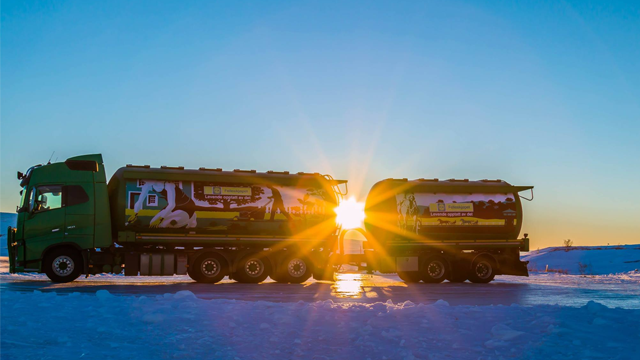 To lastebiler med grønne tanker står på snøen, med soloppgang i bakgrunnen som skaper stråler av lys.