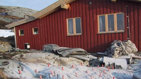 Rødt hus med vinduer og tak, omgitt av stein og snø, med rør og markeringer i bakken.