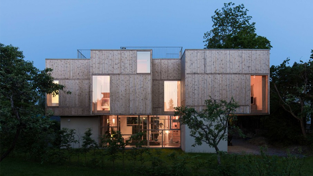 Moderne trehus med store vinduer, beliggende i en hage ved skumringstid.