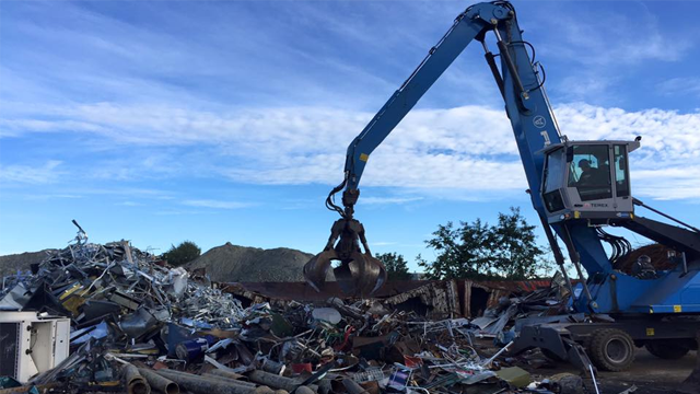 En stor kran plukker opp metallskrap på en søppelfylling under en blå himmel med skyer.
