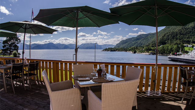 Utsikt fra en restaurantterrasse med parasoller, bord og stoler, mot en vakker innsjø og fjell.