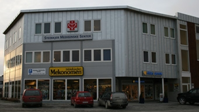 Bygningen til Steinkjer Medisinske Senter med butikker og parkerte biler foran.