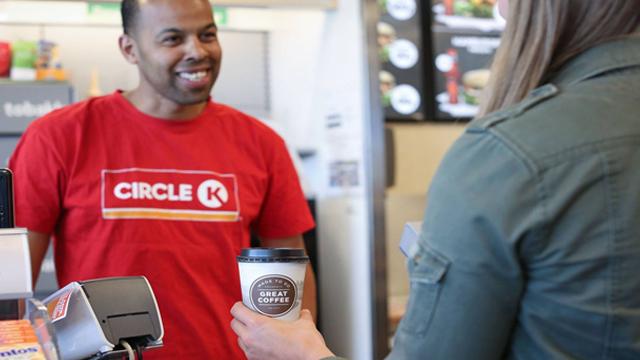 En ansatt i rød Circle K-trøye smiler mens han betjener en kunde med en kaffekopp i hånden.