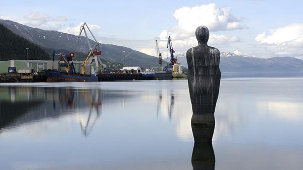 En statue står ved vannkanten med speilbilde i vannet, mens havnen og fjellene er synlige i bakgrunnen.