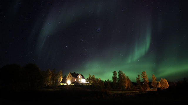 Nordlys over et hus med lys, omringet av trær under en stjerneklar natt.