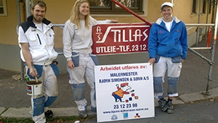 Tre malere står foran et skilt med informasjon om utleie og kontaktopplysninger for malerfirmaet.