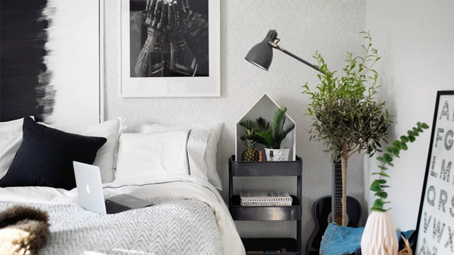 Soverom med en seng, skrivebord med laptop, planter, lampe og kunstverk på veggen.