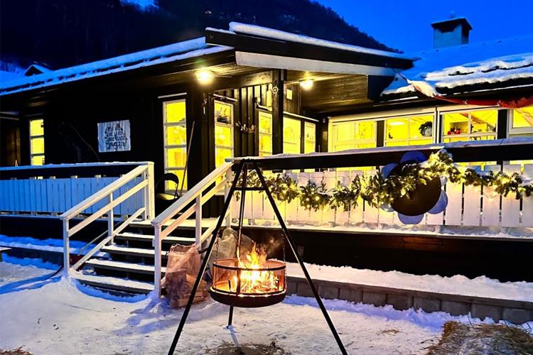 Hus med snø og bålplass, opplyst av varme lys, i en vinterlig setting med trær i bakgrunnen.
