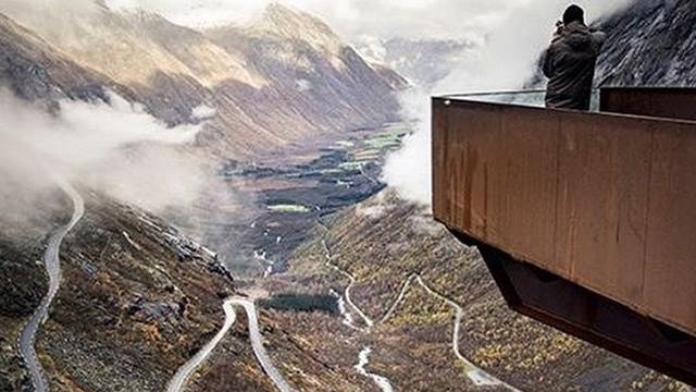 En person står på en utsiktsplattform og tar bilder av en dal med svingete veier og fjell i bakgrunnen.