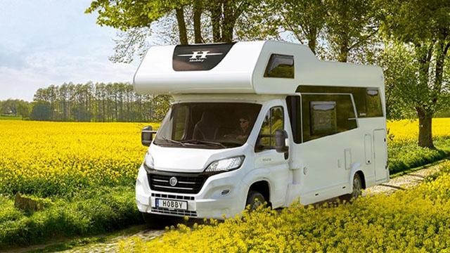 En hvit campingbil kjører på en grusvei omgitt av gule blomster og trær i bakgrunnen.