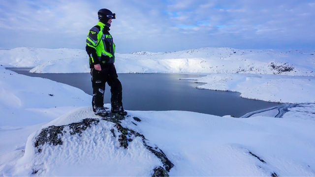 En person i en grønn og svart vinterjakke står på en snødekt stein med utsikt over en innsjø og snødekte fjell.