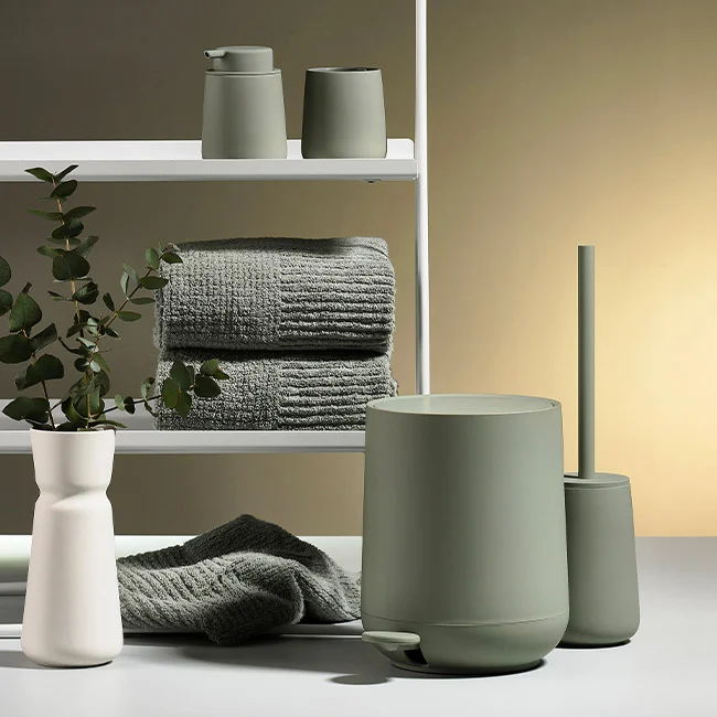 Moderne baderomsinnredning med grønne keramikkbeholdere, håndklær og en vase med stilk.