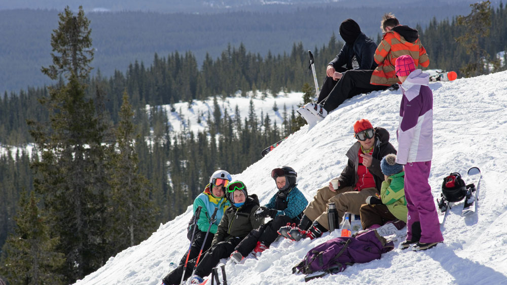 En gruppe mennesker hviler på en snødekt bakke, iført skiutstyr, med trær og fjell i bakgrunnen.