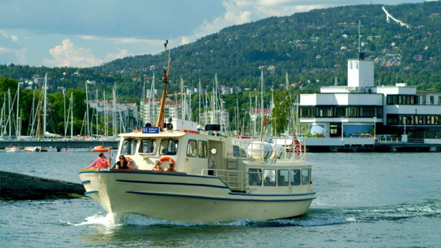 En båt seiler inn i havnen med en moderne bygning og fjell i bakgrunnen, omringet av seilbåter.