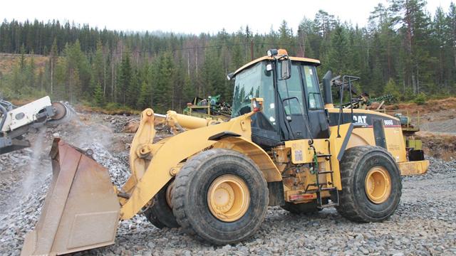 En stor gravemaskin i en steinbrudd med skog i bakgrunnen. Maskinen har en gul farge og kraftige dekk.