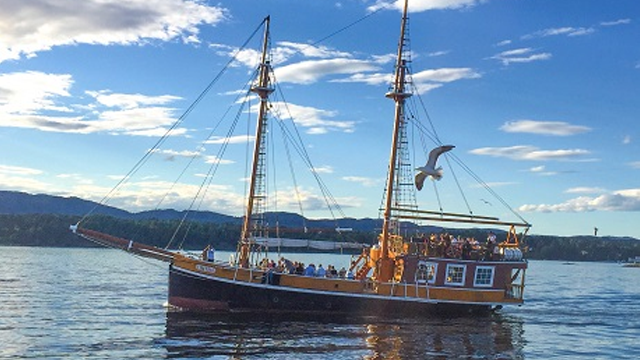 Et tradisjonelt seilskip med to master seiler på en rolig innsjø under en blå himmel med skyer.