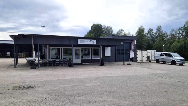 Moderne forretningsbygg med store vinduer, parkeringsplass og varebil utenfor. Grøntområde i bakgrunnen.