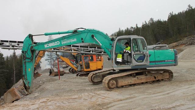 Grønn gravemaskin i arbeid på en byggeplass med flere maskiner og skog i bakgrunnen.