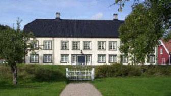 Et stort, hvitt hus med svart tak og flere vinduer, omgitt av grønt landskap og en hvit port.