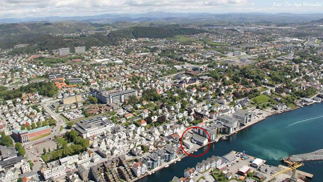 Luftbilde av en by med hvite hus og en havn, omgitt av grønne åser og blå himmel.