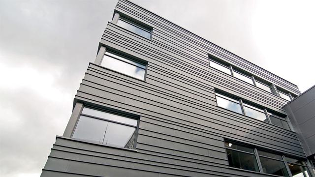 Moderne kontorbygg med grå fasade og store vinduer under en skyet himmel.