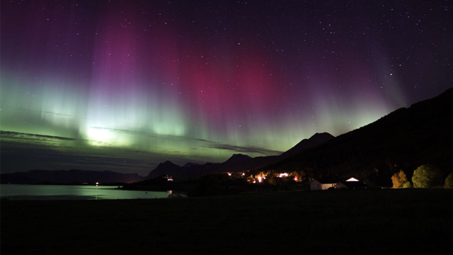 Nordlys over fjell og en liten landsby ved vannet, med farger i grønt og rosa på nattehimmelen.