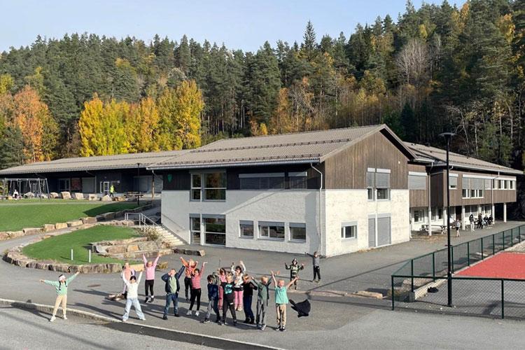 Gruppe barn og voksne som feirer foran en moderne skolebygning om høsten, med trær i bakgrunnen.