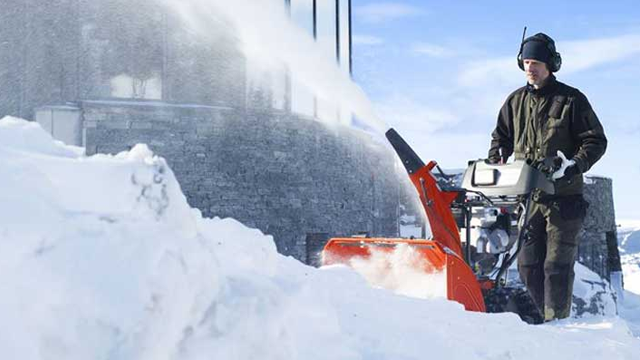 En mann bruker en snøfreser til å rydde snø foran en bygning på en solfylt vinterdag.