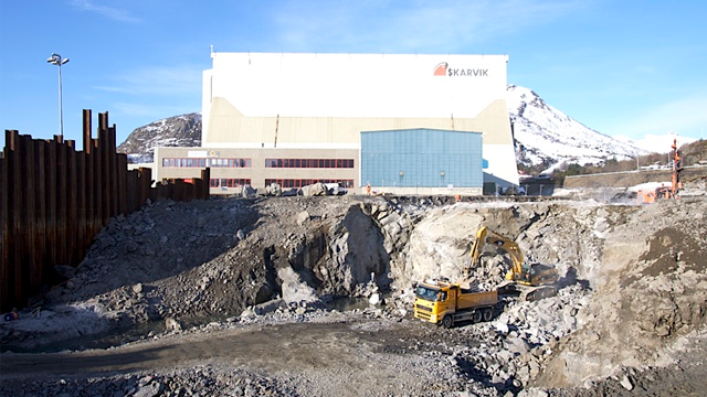 Byggeplass med gravemaskin og lastebil, med en stor fabrikkbygning i bakgrunnen og fjell i horisonten.