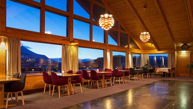En koselig restaurant med store vinduer, moderne lamper og utsikt over fjellene i skumringen.