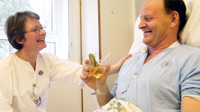 Sykepleier smiler mens hun serverer en drink til en pasient i sykesengen. Begge ser glade ut.