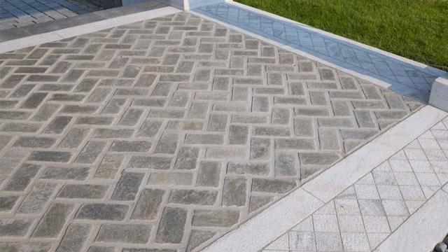 Et mønster av steinlagt terrasse med grå og beige fliser i en fiskebensformasjon. Grønne gressområder i bakgrunnen.