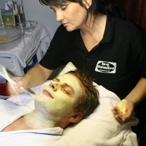 En kvinne gir en ansiktsbehandling til en mann med grønn maske på ansiktet, i et spa-miljø.