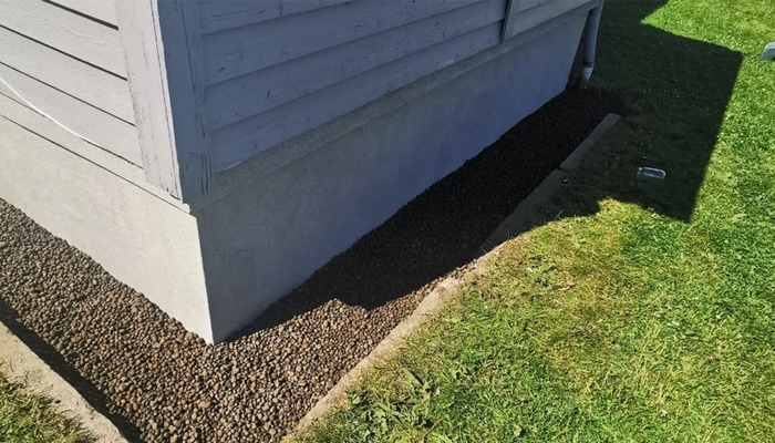Hjørnet av et hus med grå vegger, dekket av små steiner og gress i forgrunnen.