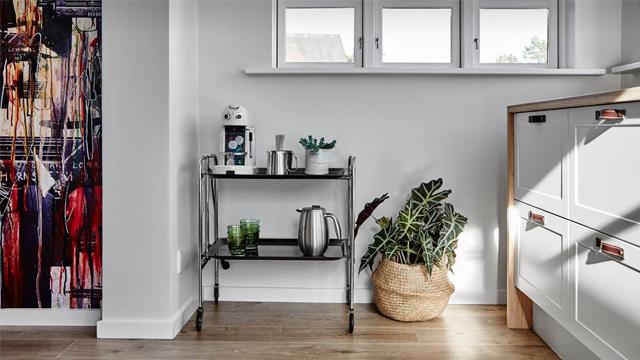 Kjøkkenhjørne med kaffemaskin, planter og en metallvogn ved vinduet, samt en hvit kjøkkenskap.