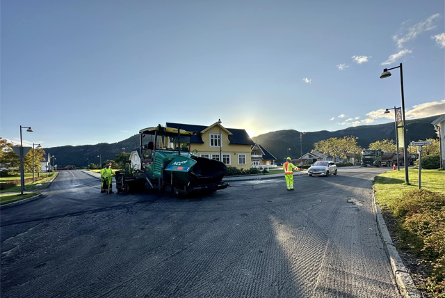 Veiarbeid med asfaltmaskin og arbeidere i gule vester ved en vei i solfylt landskap.