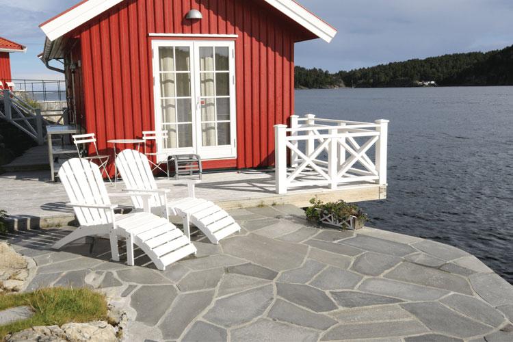 Rødt hus ved vannet med hvite stoler og steinlagt terrasse i solfylt vær.