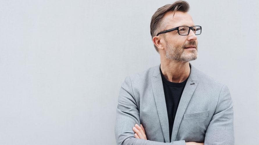 Mann med briller og grå blazer står med armene i kors mot en lys vegg.