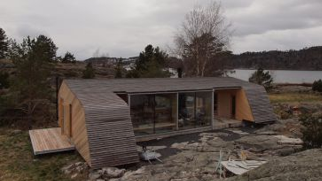 Moderne hytte med skråtak og store vinduer, beliggende ved sjøen omkranset av steiner og trær.