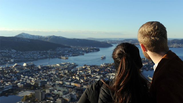 Utsikt over Bergen fra fjellet, med et par som ser utover byen og fjorden på en klar dag.