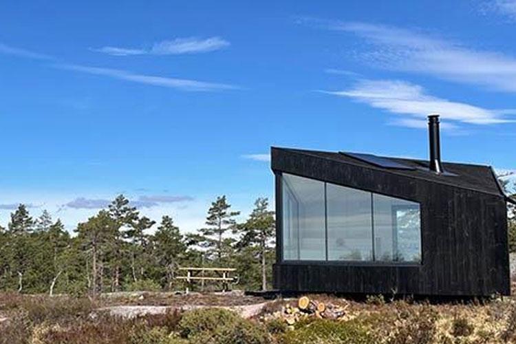Moderne hytte med store vinduer, plassert i naturen under en blå himmel med trær i bakgrunnen.